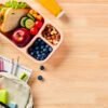 School lunch box with sandwich, fruits, vegetables, bottle of water, and kids backpack with school supplies on wooden table. Flat lay, top view.